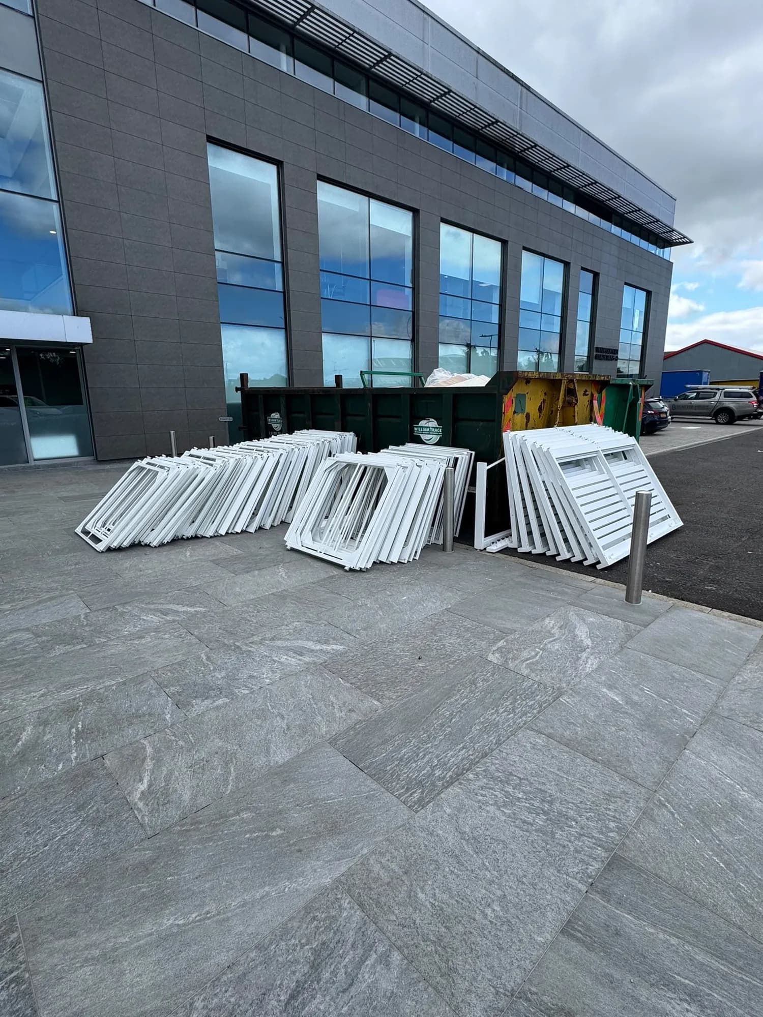 White aluminium window frames stacked on a paved forecourt beside a skip and a glass office building