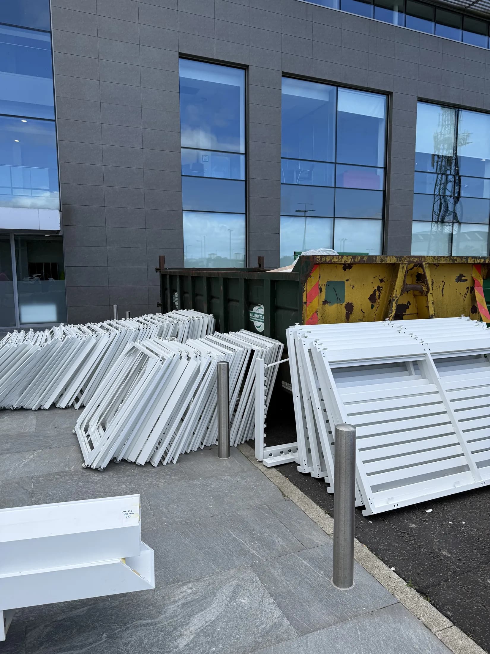 Stacks of white aluminium window frames outside a modern glass office building next to a skip