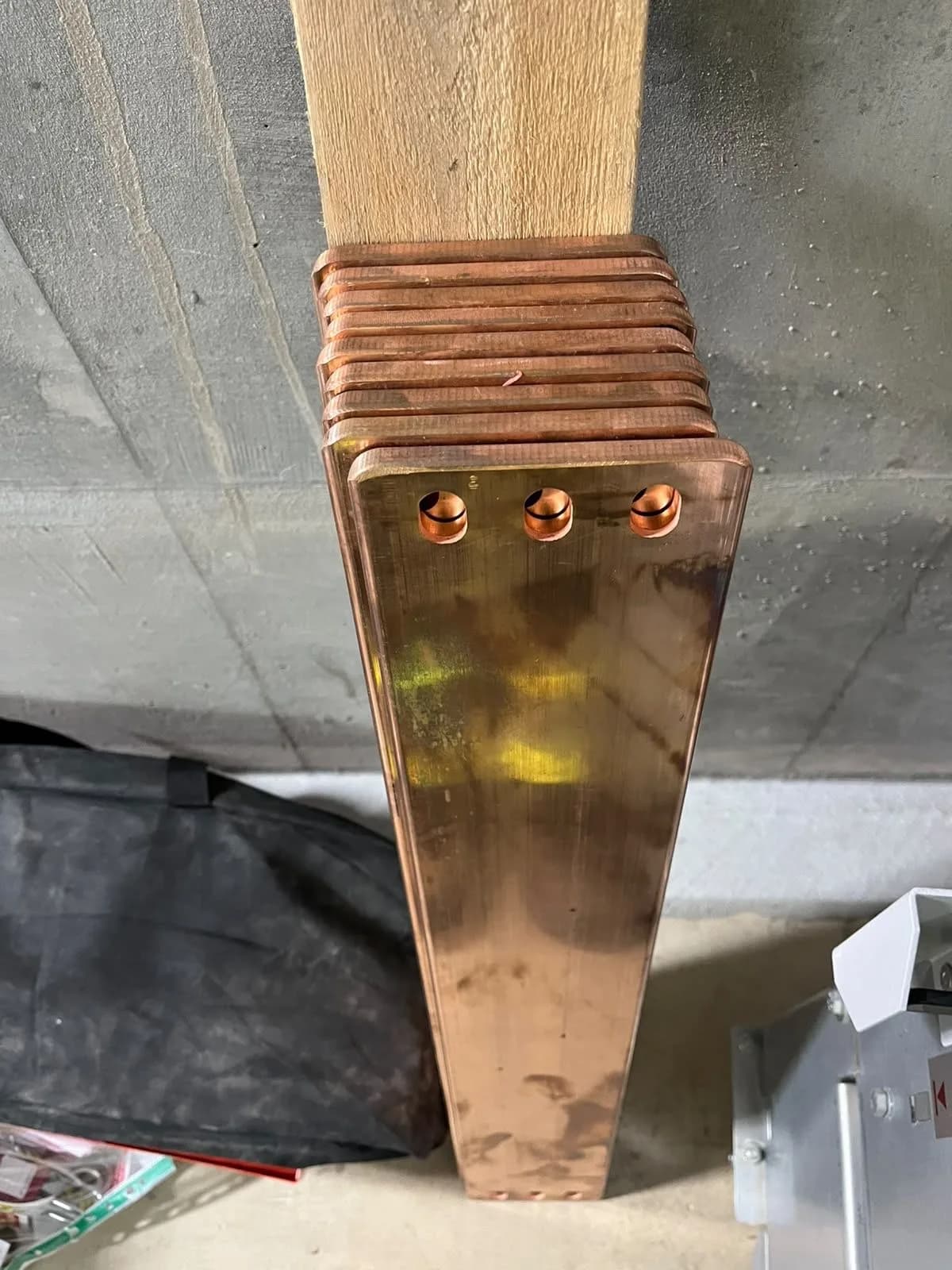 Close-up of a thick laminated copper busbar with three bolt holes, propped against a concrete wall