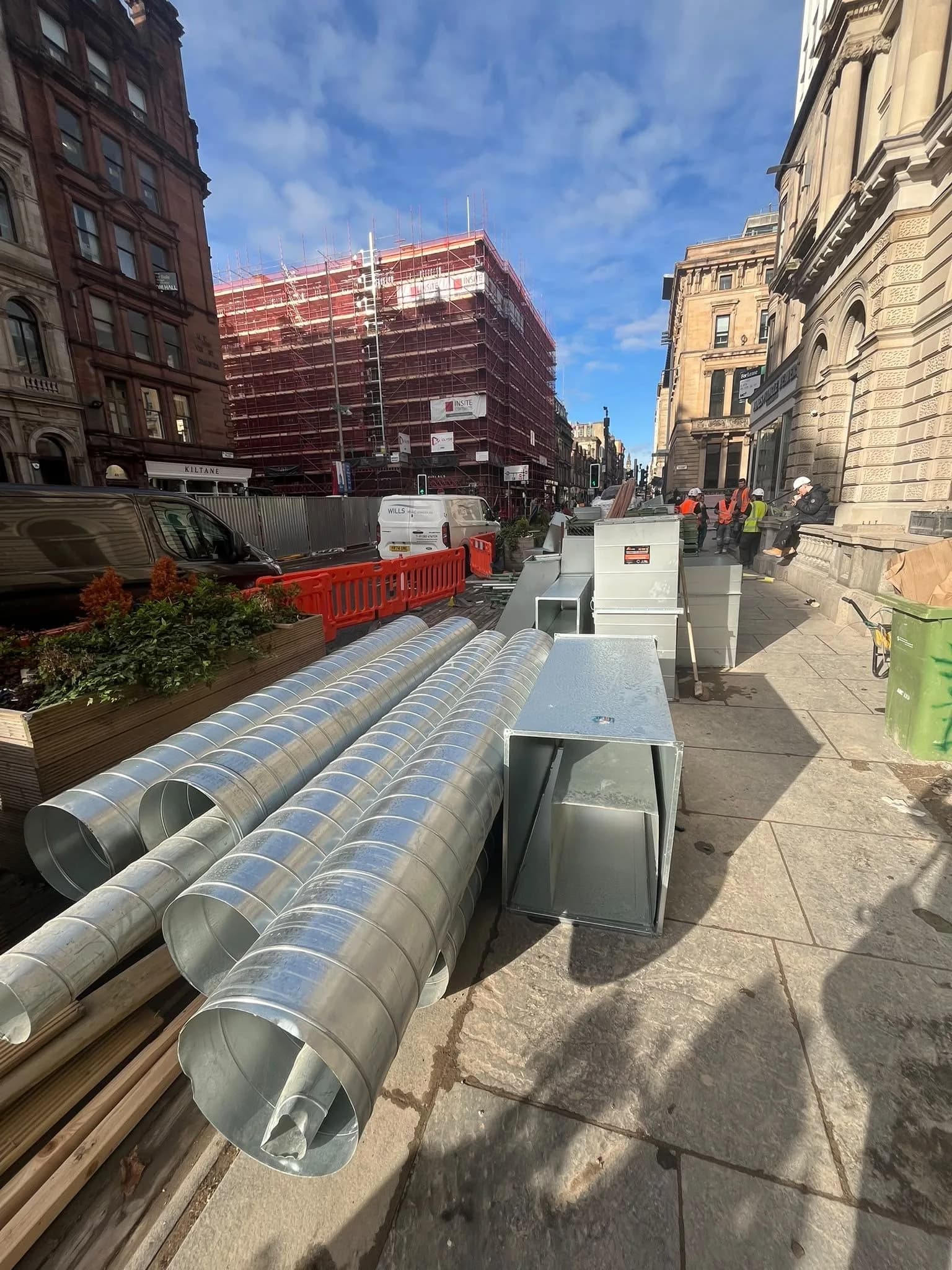 Lengths of galvanised spiral ductwork and box sections laid along a city pavement beside scaffolded sandstone buildings