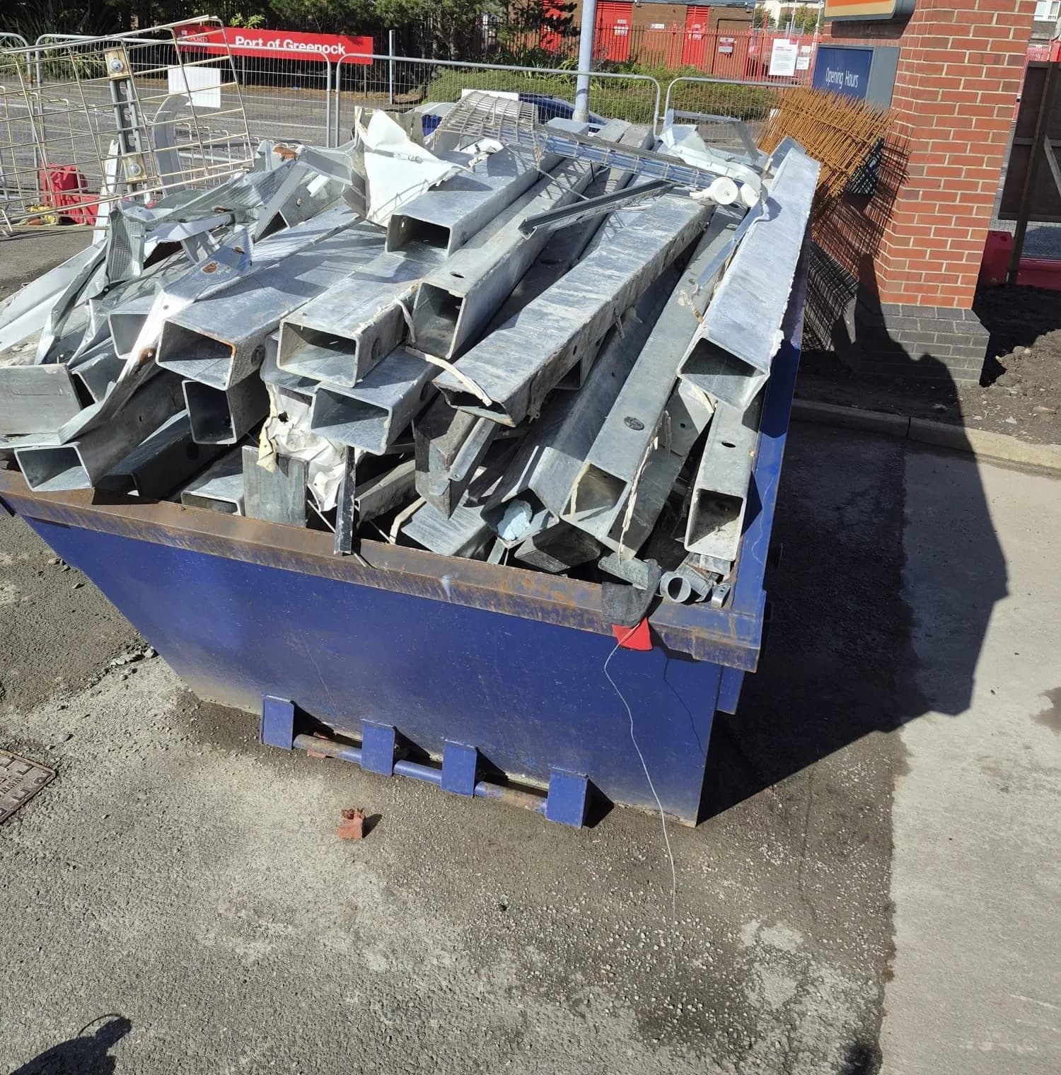 A blue skip piled high with cut galvanised steel box sections in bright sunshine