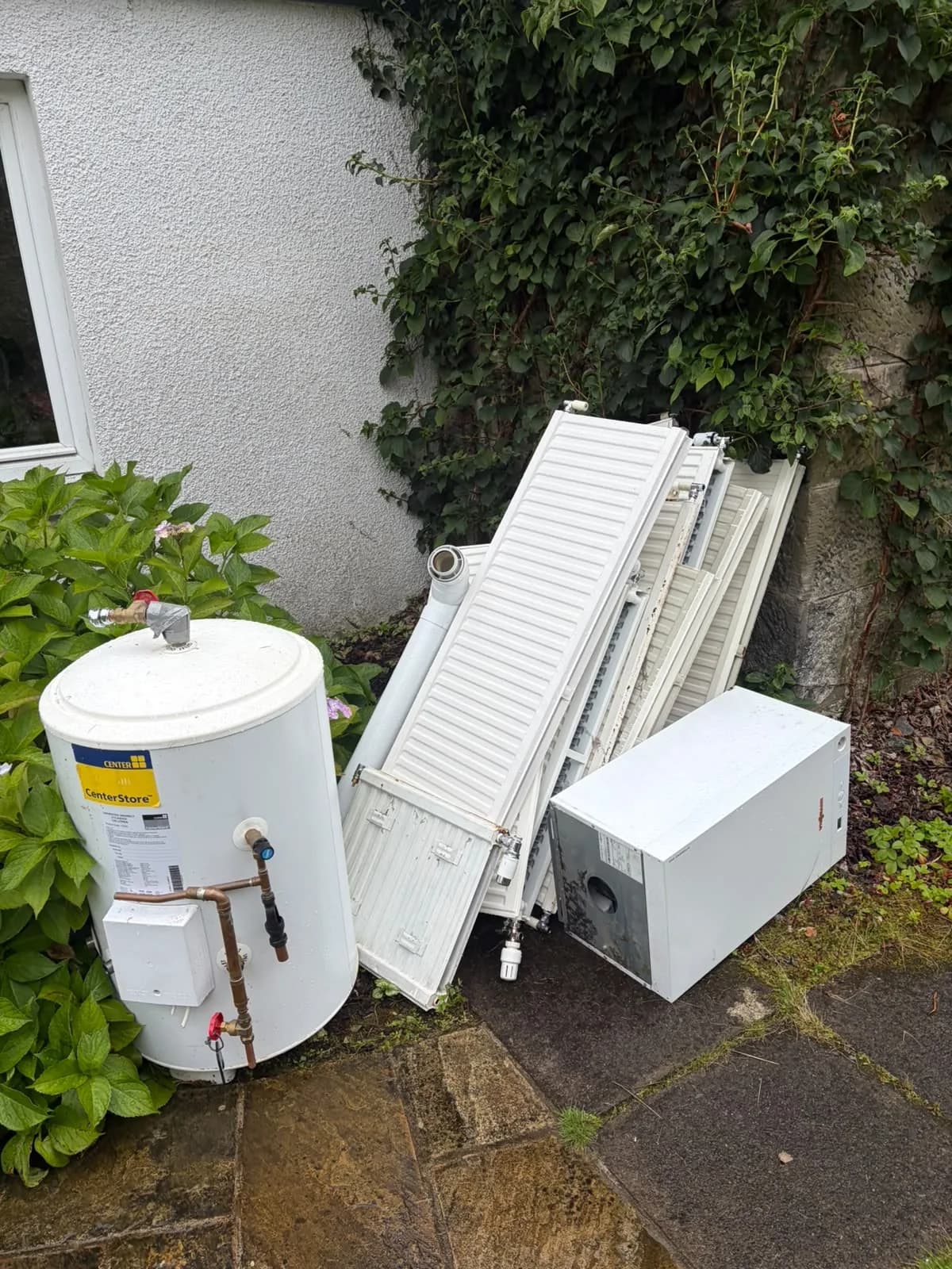 White hot water cylinder, panel radiators and a boiler unit stacked against a house wall on paving