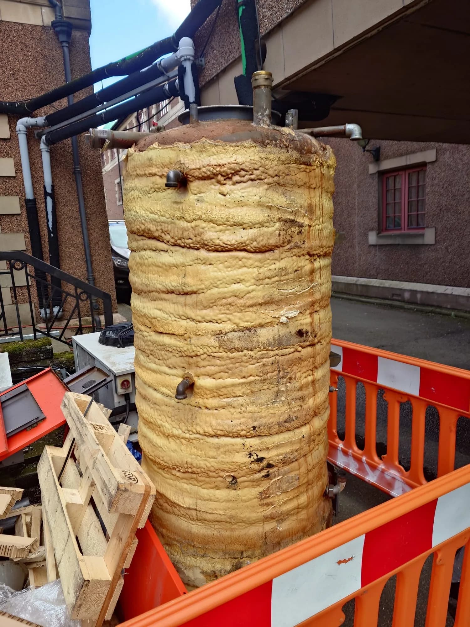 Large foam-lagged hot water cylinder inside a red and white barrier pen outside a sandstone tenement