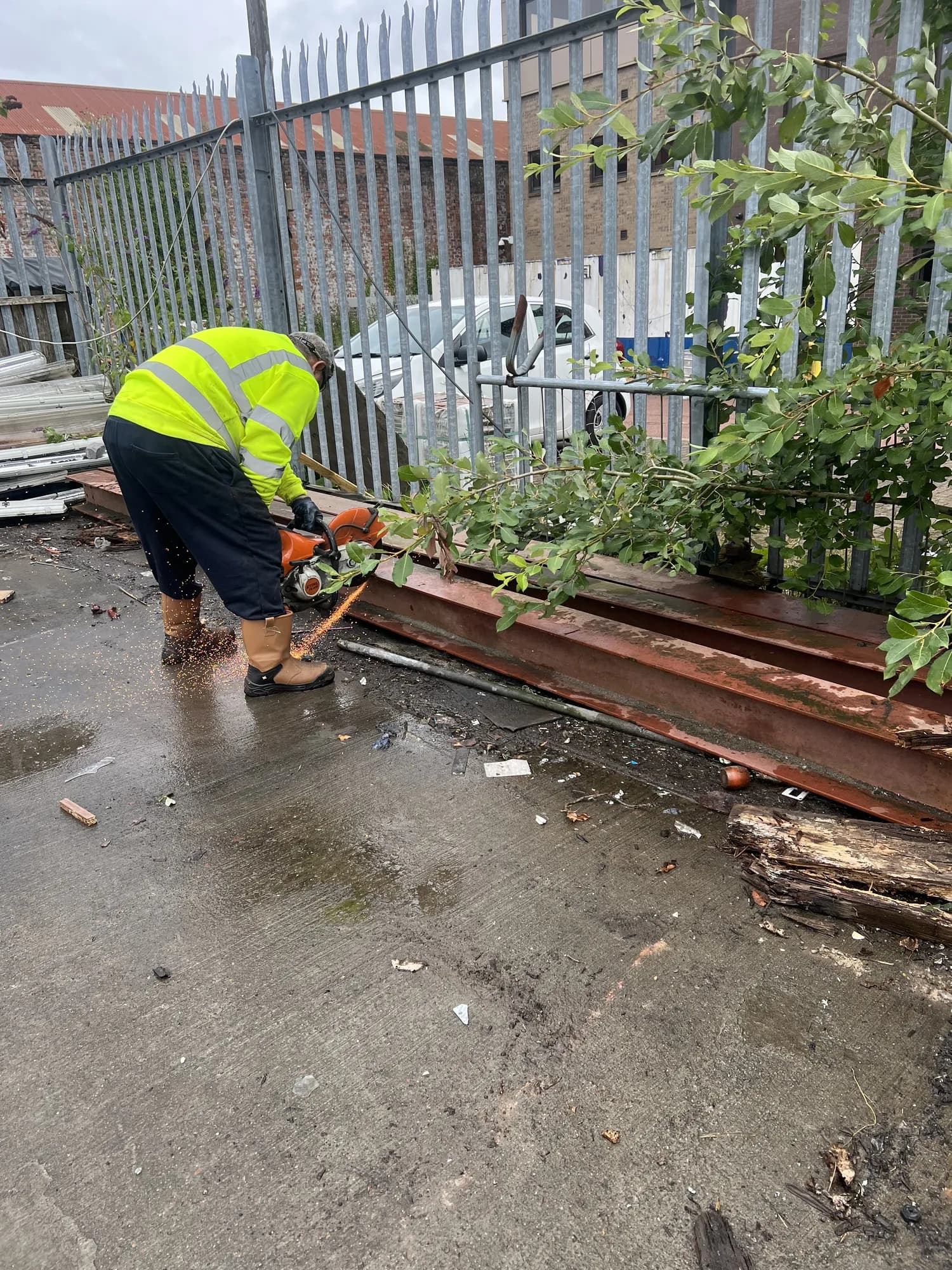 Collector in a hi-vis vest cutting rusted steel beams with a petrol saw beside a palisade fence