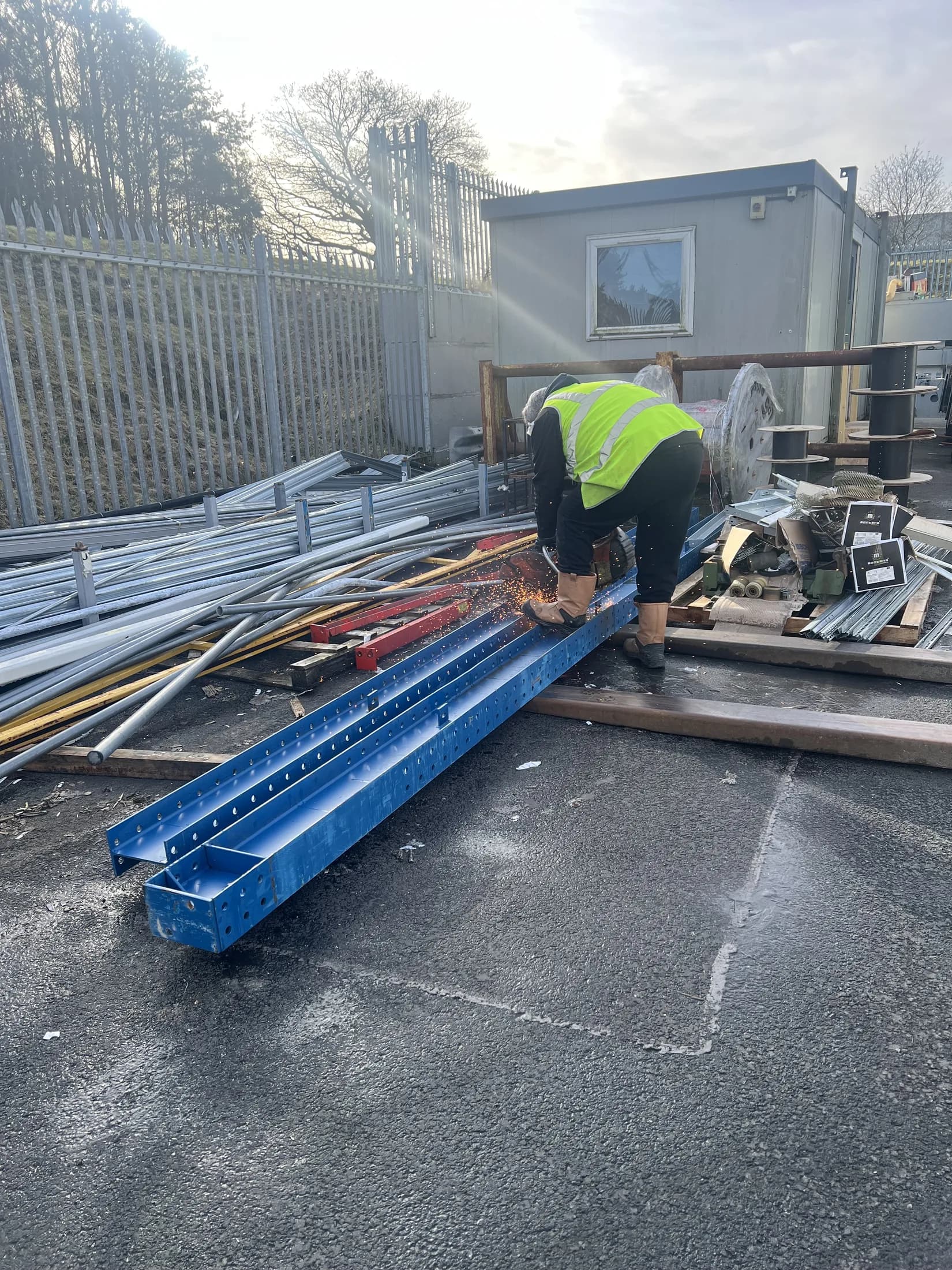 Collector in a hi-vis vest cutting blue pallet racking beams with an angle grinder in an industrial yard, sparks flying