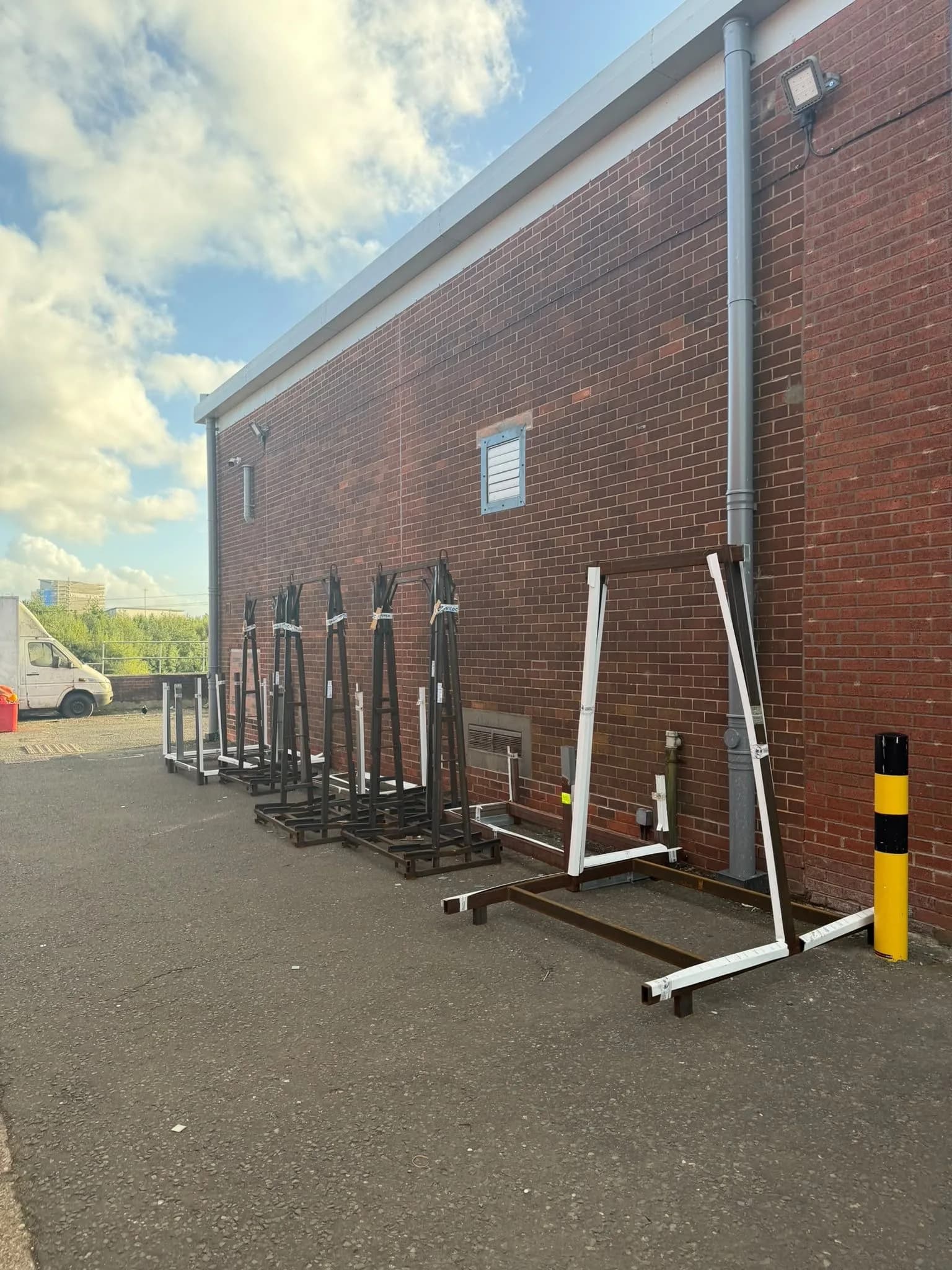 A row of steel racks and frames stacked against the outside wall of a brick building, with a white van in the background