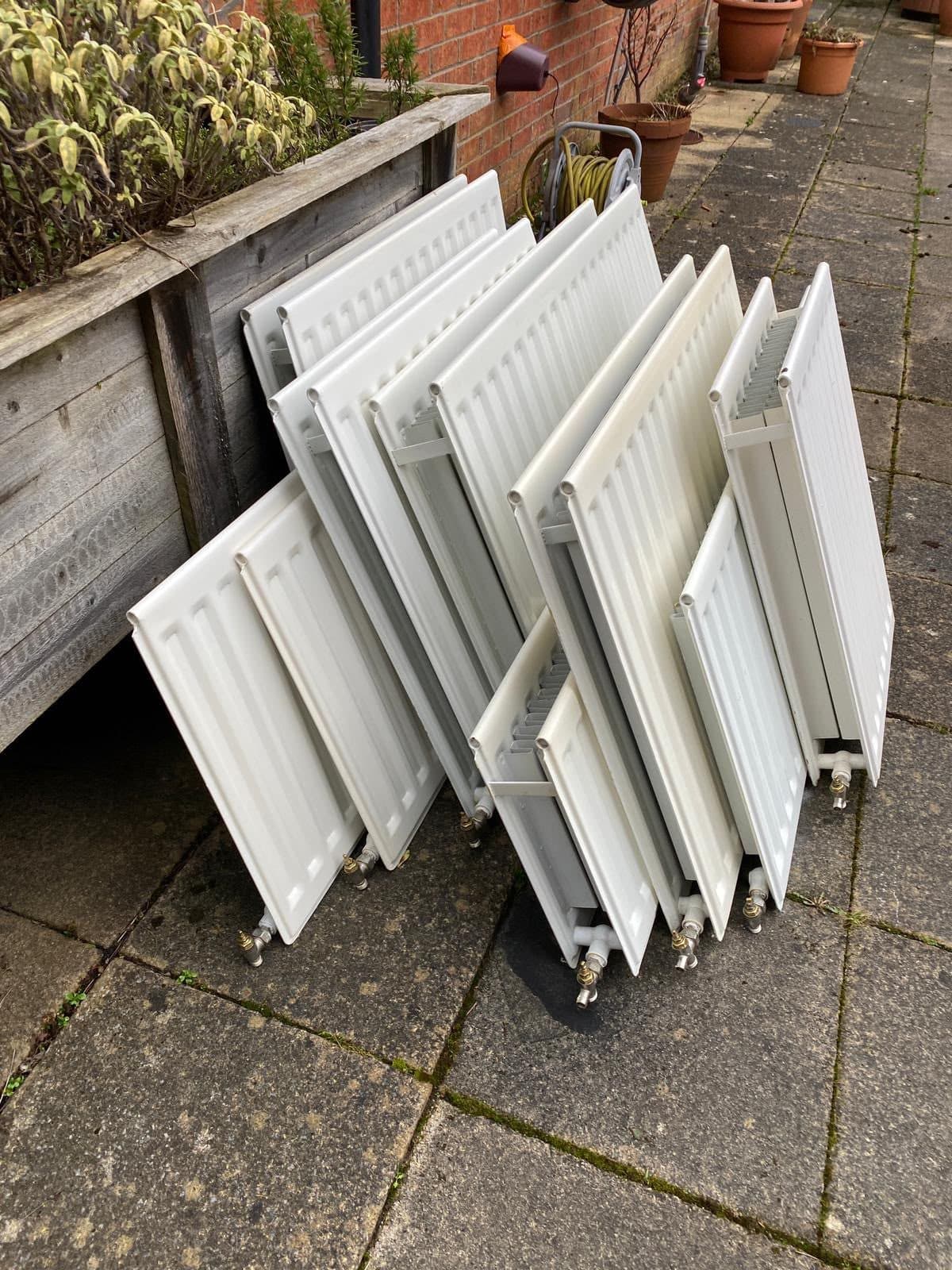 Ten white panel radiators stacked against a raised bed in a back garden, ready for collection